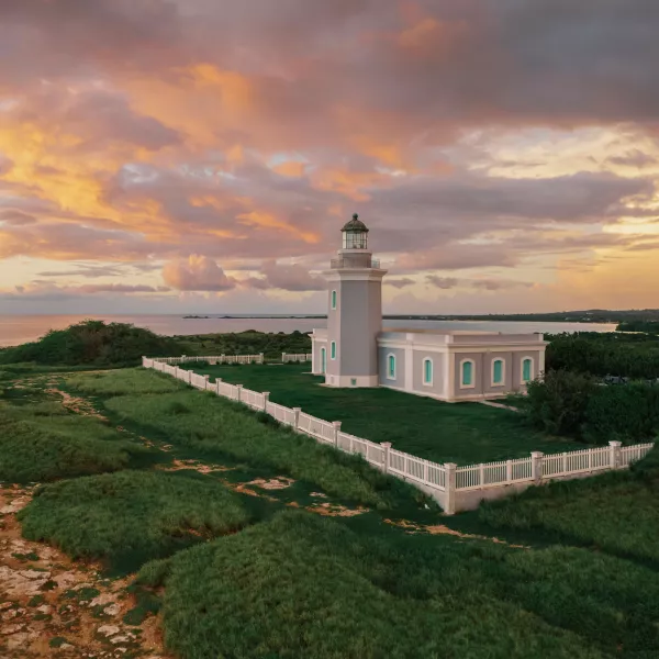 Los Morrillos Lighthouse in Cabo Rojo stands on a grassy coastal cliff, surrounded by a white fence, overlooking the ocean under a colorful sunset sky with orange and pink clouds.