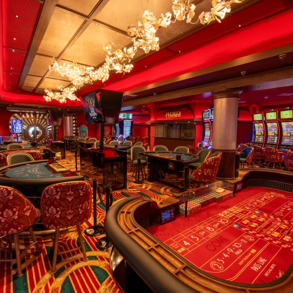 An elegant casino interior with a rich red and gold color palette, featuring a professional craps table in the foreground, blackjack tables with ornate red patterned chairs, and modern lighting fixtures hanging from a red ceiling.