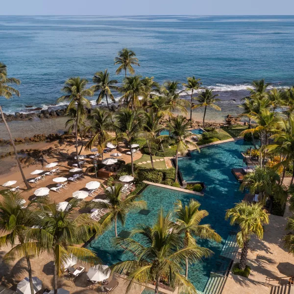 Aerial view of Dorado Beach, a Ritz-Carlton Reserve in Puerto Rico, featuring lagoon-style pools surrounded by palm trees, lounge chairs, and white umbrellas, set alongside a golden-sand beach and calm blue ocean.
