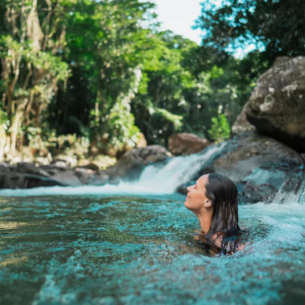 Woman swimming in a natural pool beneath a waterfall in El Yunque National Forest, surrounded by lush tropical rainforest and large rocks.