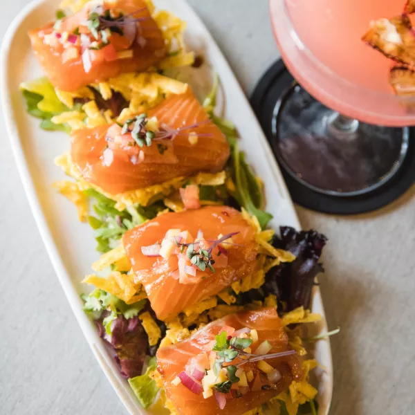 A platter of smoked salmon bites topped with fresh pico de gallo and microgreens is served alongside a pink cocktail at Lola Eclectic Cuisine in Ponce.