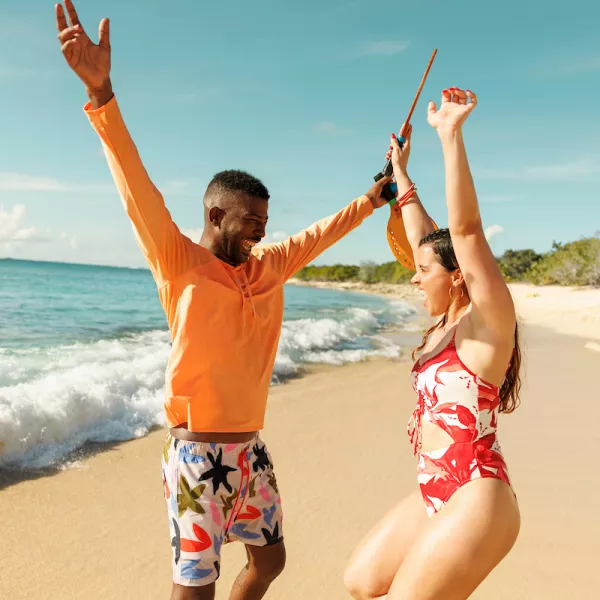 Two travelers celebrate on the shoreline of Icacos Island in Fajardo, Puerto Rico, with arms raised as waves roll onto the white-sand beach under a bright blue sky.