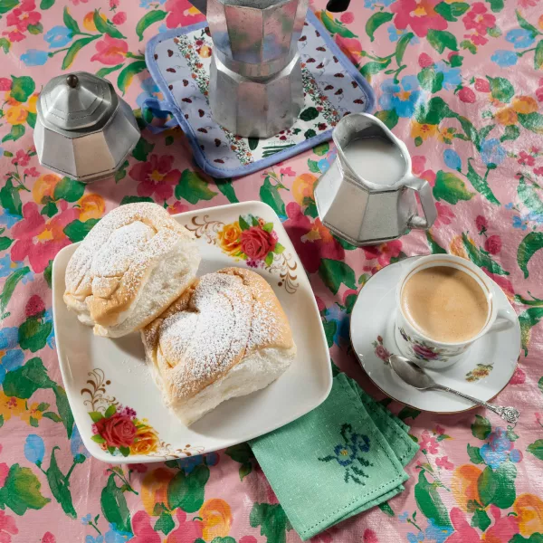 A traditional Puerto Rican breakfast featuring a cup of coffee and two mallorcas dusted with powdered sugar, served on a floral tablecloth alongside a stovetop coffee maker and creamer.