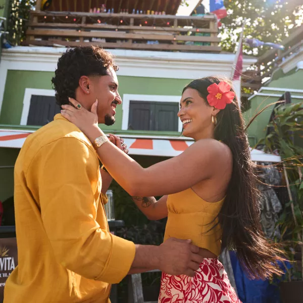 Dos personas bailan al aire libre durante una celebración festiva en Puerto Rico, con arquitectura colorida y una bandera puertorriqueña al fondo.