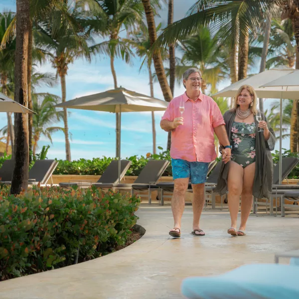 Pareja caminando por un resort tropical frente a la playa en Puerto Rico, rodeada de palmeras, sillas reclinables y vistas al océano.
