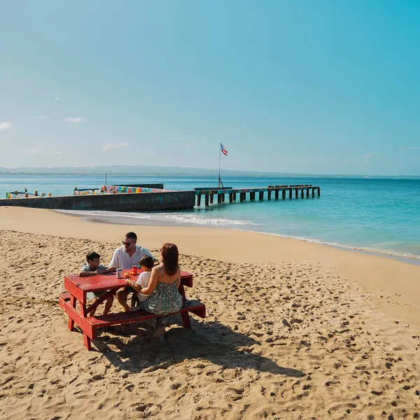 Family sitting at a picnic table on a sandy beach in Puerto Rico, with a colorful pier and Puerto Rican flag overlooking the turquoise water.