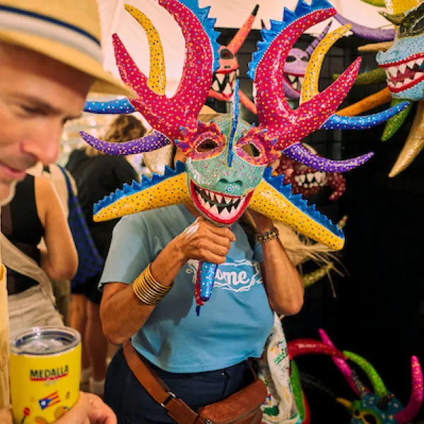 A person holds a colorful vejigante mask with long pink, yellow, and blue horns in front of their face at a booth displaying similar masks.