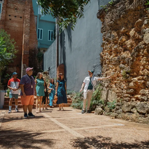 A tour guide points out historic stone ruins to a group visiting a courtyard in Old San Juan, Puerto Rico, surrounded by tropical greenery and colorful buildings.