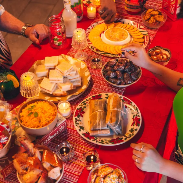 People share a Christmas spread with wrapped pasteles, rice, sandwiches, and appetizers on a red table decorated with candles.