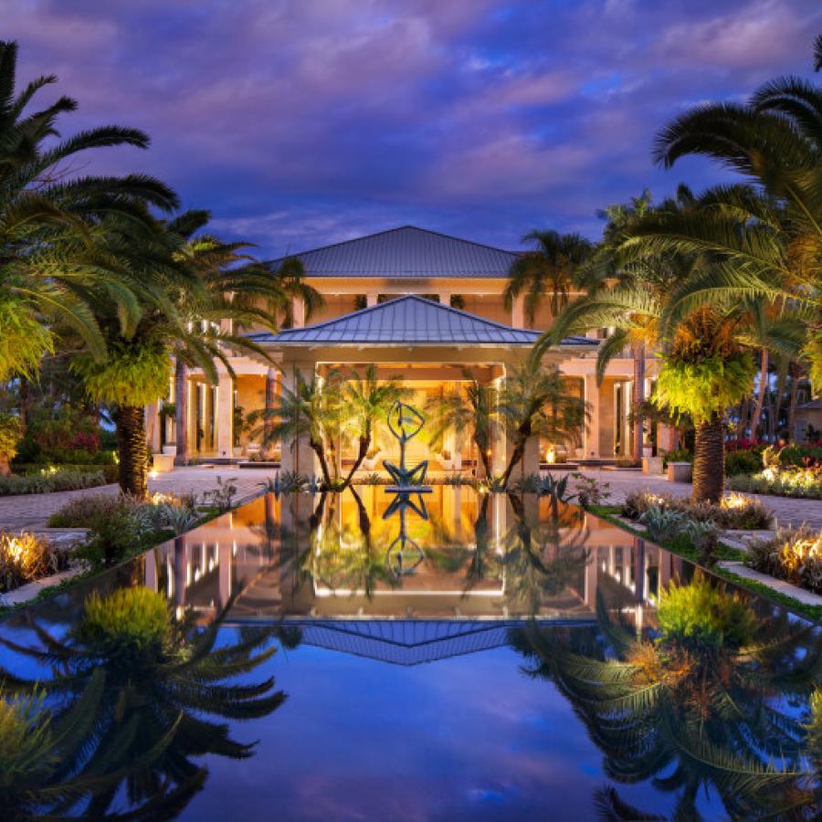 La entrada principal a un resort de lujo. Frente a la entrada hay una piscina reflectante bordeada de palmeras.