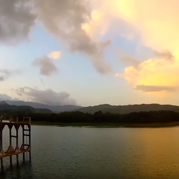 Vista panorámica de un lago y un puente al atardecer en Ponce, rodeados por montañas y un cielo iluminado por tonos dorados y nubes.