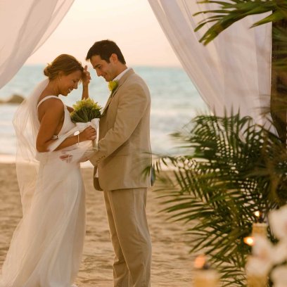A bride and groom face each other in front of an altar draped with white fabric on a beach in Puerto Rico.
