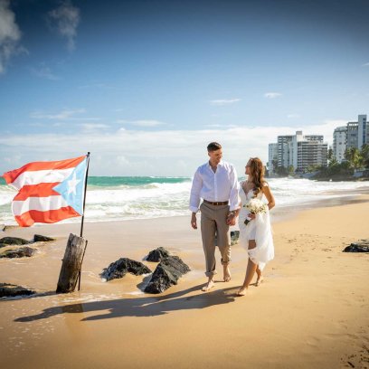 A bride and groom walk on the beach near the San Juan Marriott Resort & Stellaris Casino, with a Puerto Rican flag planted in the sand.