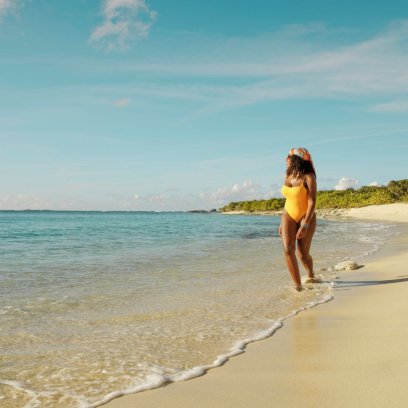 Woman in a yellow swimsuit walking along a quiet sandy beach in Puerto Rico, with gentle waves washing ashore under a clear blue sky.
