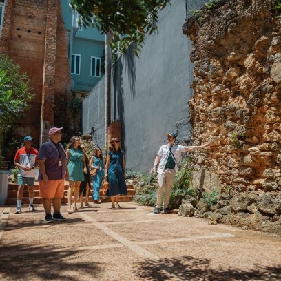 A tour guide points out historic stone ruins to a group visiting a courtyard in Old San Juan, Puerto Rico, surrounded by tropical greenery and colorful buildings.