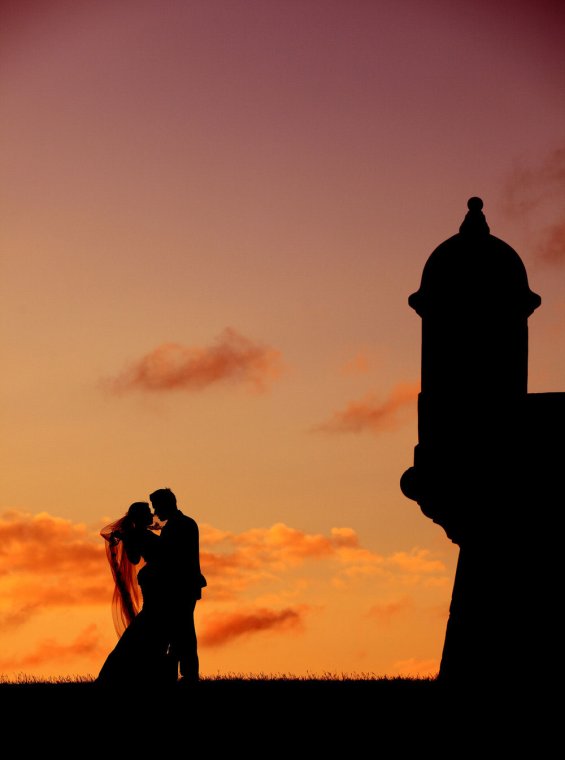 A silhouetted bride and groom embrace in front of a dramatic sunset with a historic garita in the background. San Juan, Puerto Rico.