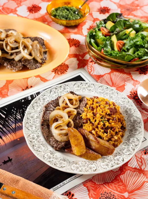 A plate of bistec encebollado topped with sautéed onions is served with arroz mamposteao and sweet fried plantains, with a fresh salad and chimichurri sauce on the side.