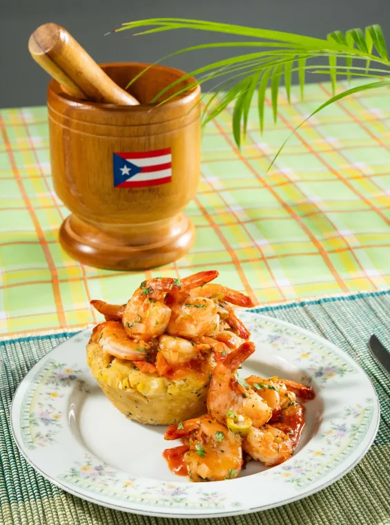 Puerto Rican mofongo topped with sautéed shrimp in sauce, served on a floral plate beside a wooden pilón decorated with the Puerto Rican flag.