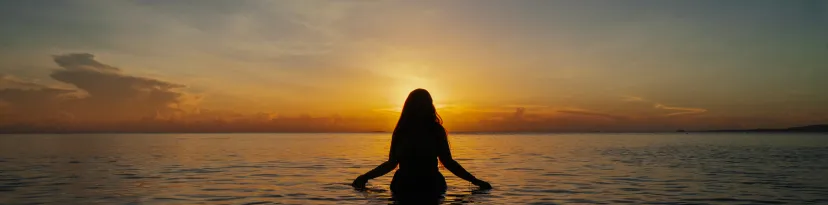 A woman stands sillouetted, hip-deep in the ocean while watching the sunset.